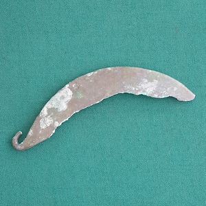 Puede incluir: Un objeto de metal de color plateado en forma de hoz con un mango curvo. El objeto está sobre un fondo de tela verde.