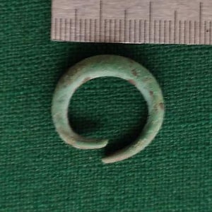 May include: A green metal ring with a diameter of approximately 2.5 cm. The ring is slightly bent and has a patina.