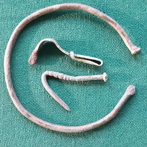 May include: An assortment of antique metal objects, including a curved ring, a small hook, and a bent nail, all with a weathered, aged appearance. The items are arranged on a textured green surface, suggesting a historical or archaeological theme.