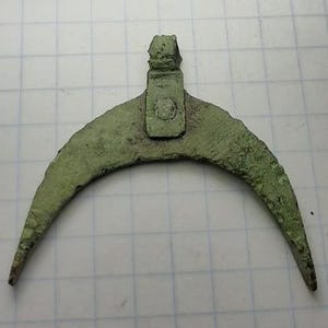 Puede incluir: Un artefacto de bronce antiguo, en forma de media luna, con un accesorio rectangular. El objeto tiene una pátina verde desgastada, lo que sugiere antigüedad. La hoja curva tiene extremos puntiagudos. El accesorio tiene un detalle circular.