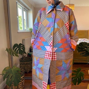 Pode incluir: Um casaco longo de patchwork em uma variedade de cores, incluindo azul, laranja, roxo e marrom. O casaco apresenta um padrão de estrela e uma borda xadrez vermelha e branca. O casaco está abotoado e tem uma gola.