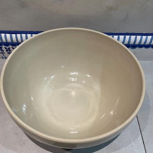 May include: A large, off-white ceramic bowl with a rounded shape. The bowl has a smooth, glossy interior and a slightly textured exterior. The neutral color and simple design make it suitable for various uses, such as serving food or as a decorative piece.