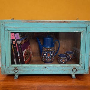 Peut inclure: Un meuble en bois turquoise avec portes vitrées, présentant une théière bleue et deux tasses assorties avec des motifs floraux. Des livres sont visibles à gauche. Le meuble repose sur de petits pieds, avec des poignées en anneau en métal.