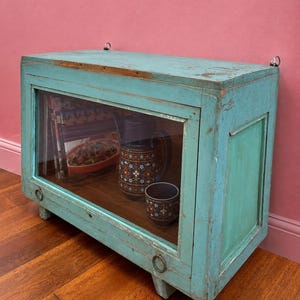 Op de afbeelding: Een rechthoekige turquoise houten vitrinekast met een glazen voorpaneel, twee metalen ringgrepen op het onderste frame en twee metalen lussen aan de bovenrand. Binnenin zijn een donkere keramische kan met patroon en een bijpassende mok zichtbaar.