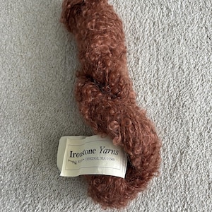 May include: A skein of brown yarn with a curly texture. A white tag with the text "Irontone Yarns" is attached. The yarn is resting on a light gray, textured surface.