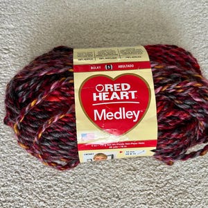 May include: A skein of Red Heart Medley yarn in shades of red, burgundy, gray, and yellow. The label features a red heart logo and the text "Medley". The yarn is labeled as bulky, weighing 4 oz (113 g) and 66 yds (60 m).