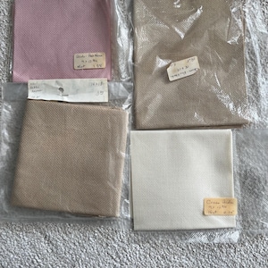 May include: Four pieces of cross-stitch fabric in various colors: light pink, beige, tan, and white. Each piece is packaged in a clear plastic bag with a handwritten label. The fabric sizes vary, with one piece measuring 14 x 18 inches.