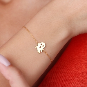 Könnte beinhalten: Ein goldfarbenes Armband mit einem kleinen Geisteranhänger. Der Anhänger hat ausgeschnittene Augen und einen Mund. Das Armband wird an einem Handgelenk vor einem roten Oberteil getragen. Der Hintergrund ist eine schlichte weiße Wand.