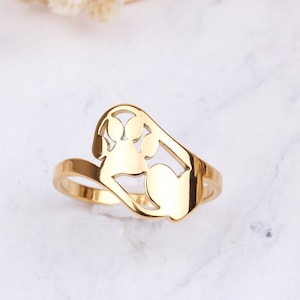 May include: A gold-colored ring with a paw print and heart design. The ring is set against a white marble surface with dried flowers. The ring's design features a cut-out paw print and a heart shape.