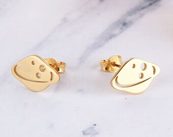 Puede incluir: Un par de pendientes de color dorado con forma de planeta con un anillo alrededor. Los pendientes tienen un diseño simple y minimalista.