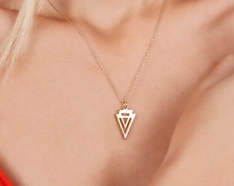 May include: Gold-colored necklace with a geometric pendant. The pendant features a layered triangle design. The necklace is worn against a red top. The chain is delicate and the pendant is centered.