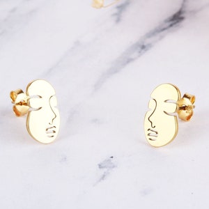 May include: A pair of gold-toned earrings featuring a minimalist face design. The earrings are small and delicate, perfect for everyday wear.