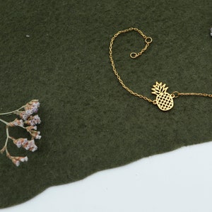 Puede incluir: Un brazalete de cadena dorada con un dije de piña. El brazalete está sobre un fondo de tela verde.