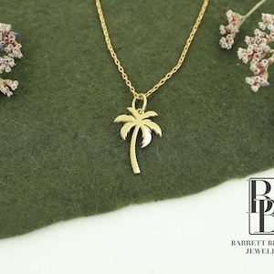 May include: A gold palm tree pendant necklace on a green background. The necklace is delicate and features a simple palm tree design. The pendant is about 1 inch tall.