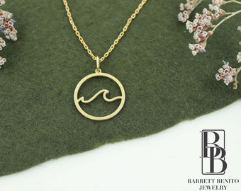 May include: Gold necklace with a circle pendant featuring a wave design. The necklace is on a green fabric background.