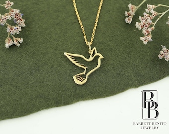May include: A gold necklace with a delicate, cutout dove pendant. The dove is a symbol of peace and hope. The necklace is on a green background.