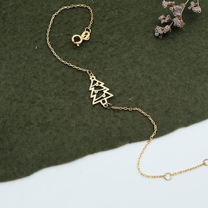 Puede incluir: Una delicada pulsera dorada con un dije de árbol de Navidad. El dije es un árbol estilizado con tres niveles. La pulsera está sobre una superficie verde oscuro con flores secas.