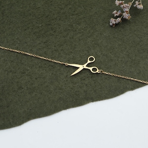 May include: A gold-colored bracelet featuring a scissors charm. The scissors are in a side profile, with the blades slightly open. The bracelet chain is delicate and thin. The background is a dark green fabric with dried flowers.
