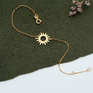 Könnte beinhalten: Ein goldenes Kettenarmband mit einem Sonnenanhänger. Das Armband liegt auf einem grünen Stoffuntergrund.