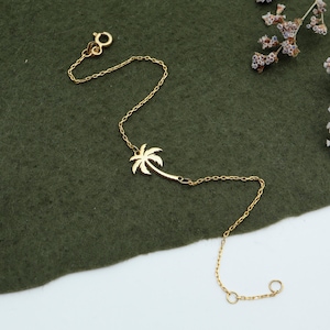 May include: A gold palm tree charm bracelet with a delicate chain. The bracelet is on a green fabric background.