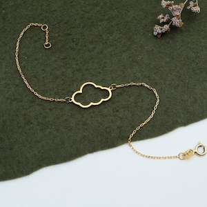 May include: A delicate gold bracelet with a cloud-shaped charm. The bracelet has a fine chain and a lobster clasp. The jewellery is displayed on a dark green surface with dried flowers.