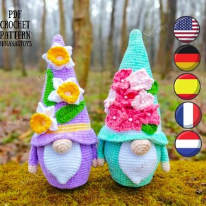 May include: Two crocheted gnomes with floral hats. One gnome has a purple hat with yellow and white daffodils. The other gnome has a turquoise hat with pink flowers. The gnomes are sitting on a mossy log. The image includes the text "PDF CROCHET PATTERN ANNANASTOYS".
