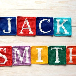 Może przedstawiać: Połączone kwadraty na szydełku, tworzące imię "JACK SMITH" białymi literami na r&oacute;żnokolorowych tłach. Kwadraty są ułożone w linii na jasnym drewnianym tle.