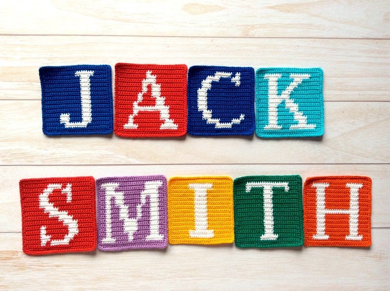 Może przedstawiać: Połączone kwadraty na szydełku, tworzące imię "JACK SMITH" białymi literami na r&oacute;żnokolorowych tłach. Kwadraty są ułożone w linii na jasnym drewnianym tle.