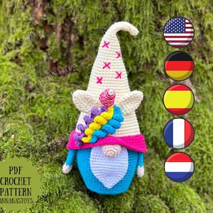 May include: A crochet gnome pattern featuring a white hat with pink and white X's, a blue body, and a rainbow-coloured beard. The pattern is available in PDF format and is designed by Anna Nas Toys.