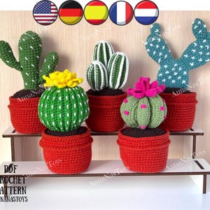 May include: A collection of handmade crochet cactus plants in red pots. The cacti vary in shape and color, including green with white accents and a yellow flower, and blue with white accents. The display includes the text "PDF CROCHET PATTERN ANNANASTOYS".