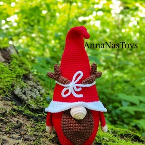 Puede incluir: Un gnomo de crochet rojo con barba blanca y un lazo blanco. El gnomo tiene cuernos y lleva un gorro rojo con ribete blanco. El gnomo est&aacute; sentado en un tronco cubierto de musgo. El texto "AnnaNasToys" es visible en el fondo.