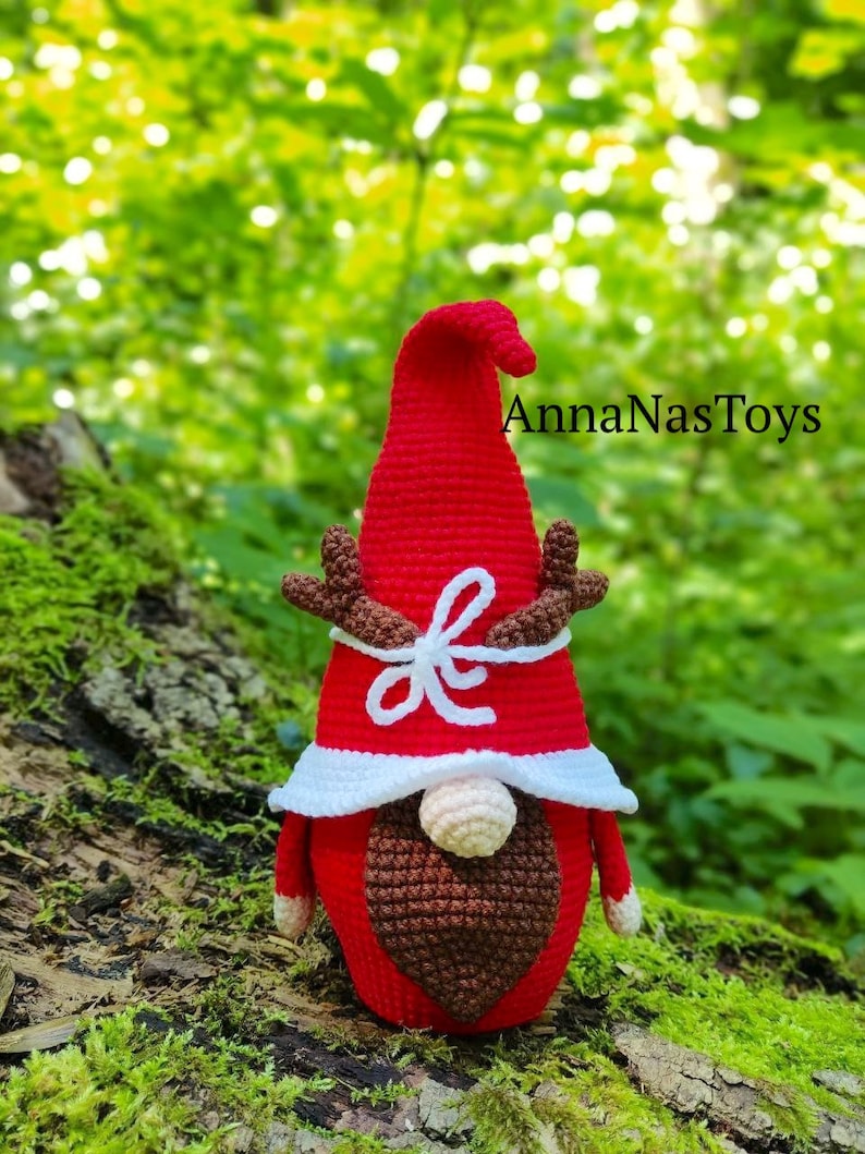 Puede incluir: Un gnomo de crochet rojo con barba blanca y un lazo blanco. El gnomo tiene cuernos y lleva un gorro rojo con ribete blanco. El gnomo est&aacute; sentado en un tronco cubierto de musgo. El texto "AnnaNasToys" es visible en el fondo.
