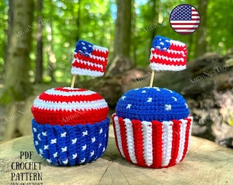 May include: Two crocheted cupcake toys decorated with red, white, and blue stripes and stars. Each cupcake has a small American flag on top. The cupcakes are sitting on a wooden surface.