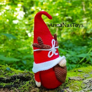 Puede incluir: Un gnomo de crochet rojo con barba blanca y cuernos marrones. El gnomo lleva un gorro de crochet blanco y est&aacute; de pie sobre un tronco cubierto de musgo. El texto "AnnaNasToys" es visible en el fondo.