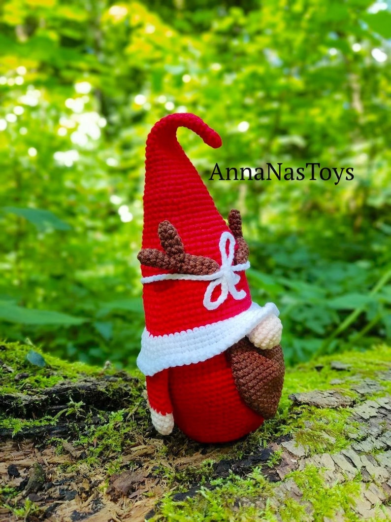 Puede incluir: Un gnomo de crochet rojo con barba blanca y cuernos marrones. El gnomo lleva un gorro de crochet blanco y est&aacute; de pie sobre un tronco cubierto de musgo. El texto "AnnaNasToys" es visible en el fondo.