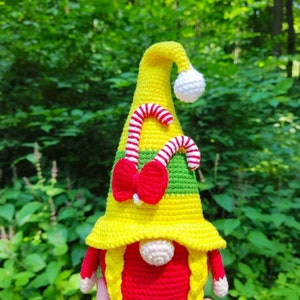 Puede incluir: Un gnomo de crochet con un sombrero amarillo, cuerpo rojo y trenzas amarillas. El sombrero tiene un dise&ntilde;o de bast&oacute;n de caramelo y un lazo rojo. El gnomo sostiene un bast&oacute;n de caramelo en su mano. El texto "AnnaNasToys" es visible en la parte inferior de la imagen.