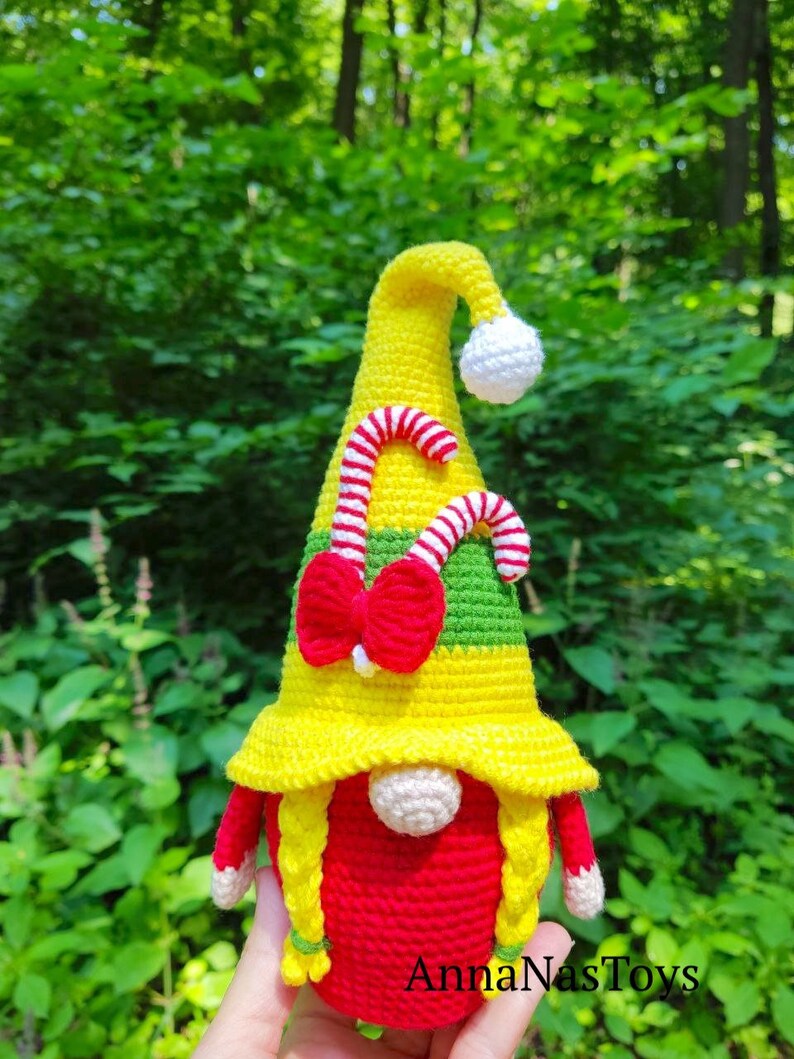 Puede incluir: Un gnomo de crochet con un sombrero amarillo, cuerpo rojo y trenzas amarillas. El sombrero tiene un dise&ntilde;o de bast&oacute;n de caramelo y un lazo rojo. El gnomo sostiene un bast&oacute;n de caramelo en su mano. El texto "AnnaNasToys" es visible en la parte inferior de la imagen.