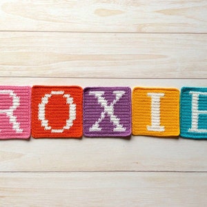 Może przedstawiać: Pięć kwadrat&oacute;w na szydełku, kt&oacute;re tworzą imię "ROXIE" białymi literami. Każdy kwadrat jest w innym kolorze: r&oacute;żowy, pomarańczowy, fioletowy, ż&oacute;łty i niebieski.