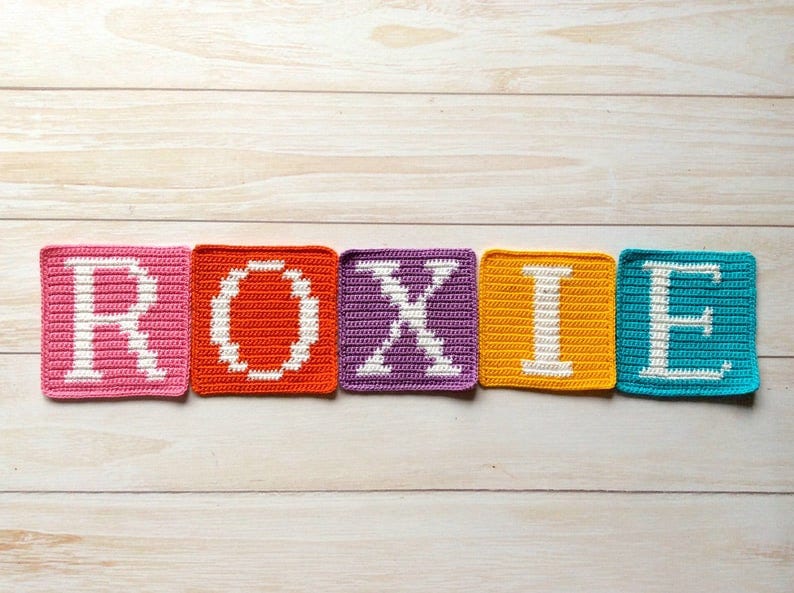 Może przedstawiać: Pięć kwadrat&oacute;w na szydełku, kt&oacute;re tworzą imię "ROXIE" białymi literami. Każdy kwadrat jest w innym kolorze: r&oacute;żowy, pomarańczowy, fioletowy, ż&oacute;łty i niebieski.