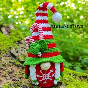 Puede incluir: Un gnomo navide&ntilde;o de crochet con un gorro a rayas rojo y blanco, un borde de gorro verde y un copo de nieve blanco en la parte delantera. El gnomo sostiene un bast&oacute;n de caramelo en su mano. El gnomo est&aacute; sentado en un tronco cubierto de musgo. El texto "AnnaNasToys" es visible en el fondo.
