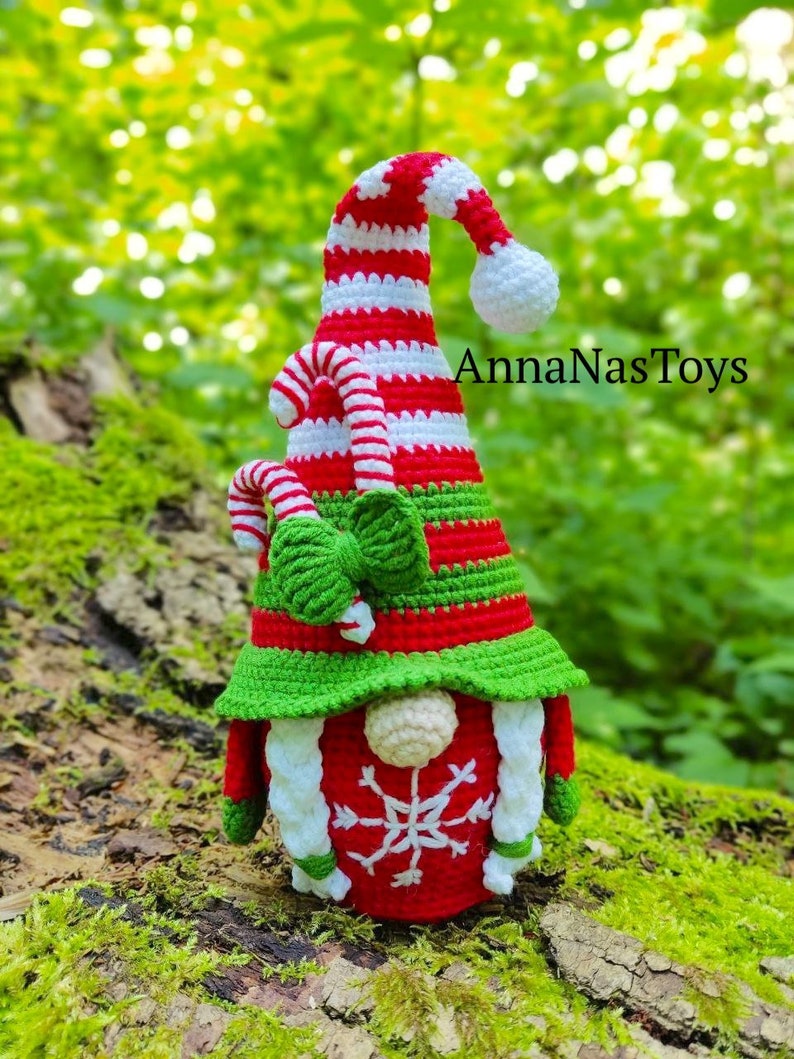 Puede incluir: Un gnomo navide&ntilde;o de crochet con un gorro a rayas rojo y blanco, un borde de gorro verde y un copo de nieve blanco en la parte delantera. El gnomo sostiene un bast&oacute;n de caramelo en su mano. El gnomo est&aacute; sentado en un tronco cubierto de musgo. El texto "AnnaNasToys" es visible en el fondo.