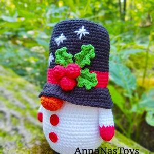 May include: A crocheted snowman toy wearing a black hat with white snowflakes and a red and green holly design. The snowman has a white body, red buttons, and a red and white striped scarf. The snowman is sitting on a mossy log. The text "AnnaNasToys" is visible at the bottom of the image.