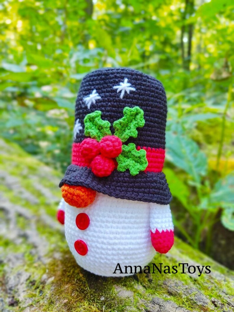May include: A crocheted snowman toy wearing a black hat with white snowflakes and a red and green holly design. The snowman has a white body, red buttons, and a red and white striped scarf. The snowman is sitting on a mossy log. The text "AnnaNasToys" is visible at the bottom of the image.
