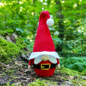 Puede incluir: Un gnomo de crochet rojo y blanco con barba blanca y cintur&oacute;n negro. El gnomo est&aacute; sentado en un tronco cubierto de musgo en un entorno de bosque. El texto "AnnaNasToys" es visible en el fondo.