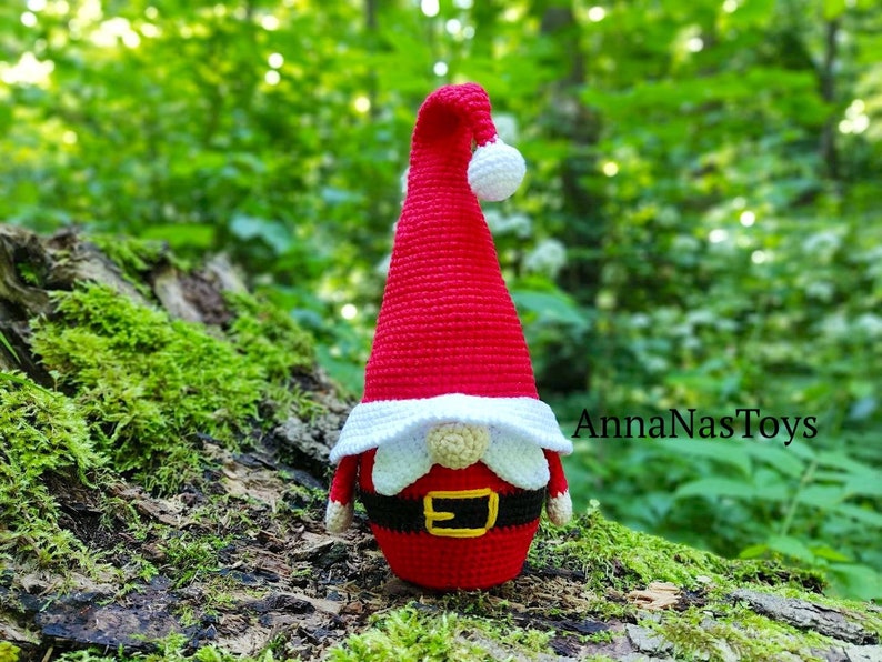 Puede incluir: Un gnomo de crochet rojo y blanco con barba blanca y cintur&oacute;n negro. El gnomo est&aacute; sentado en un tronco cubierto de musgo en un entorno de bosque. El texto "AnnaNasToys" es visible en el fondo.