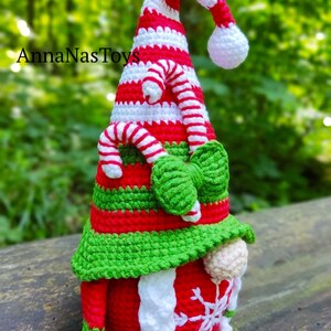 Puede incluir: Un gnomo de Navidad de crochet con un gorro a rayas rojo y blanco, barba verde y un copo de nieve blanco en el vientre. El gnomo lleva un gorro verde con un dise&ntilde;o de bast&oacute;n de caramelo.