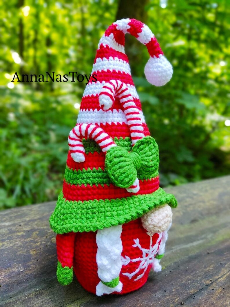 Puede incluir: Un gnomo de Navidad de crochet con un gorro a rayas rojo y blanco, barba verde y un copo de nieve blanco en el vientre. El gnomo lleva un gorro verde con un dise&ntilde;o de bast&oacute;n de caramelo.