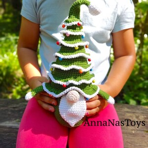 Puede incluir: Un juguete de gnomo de &aacute;rbol de Navidad de crochet con un sombrero verde, barba blanca y adornos coloridos. El gnomo est&aacute; siendo sostenido por un ni&ntilde;o que lleva una camisa azul claro y pantalones rosas. La imagen incluye el texto "AnnaNasToys".