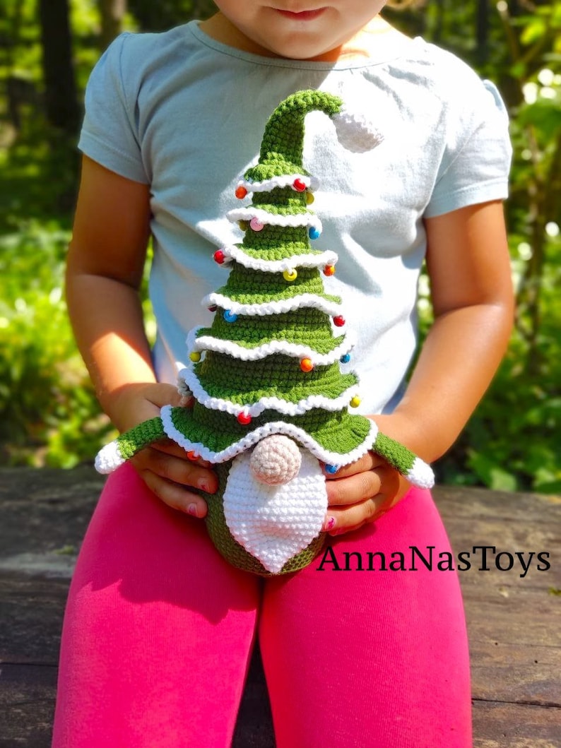 Puede incluir: Un juguete de gnomo de &aacute;rbol de Navidad de crochet con un sombrero verde, barba blanca y adornos coloridos. El gnomo est&aacute; siendo sostenido por un ni&ntilde;o que lleva una camisa azul claro y pantalones rosas. La imagen incluye el texto "AnnaNasToys".