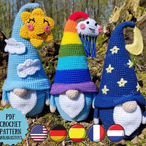 May include: Three crocheted gnomes with different hats. The first gnome has a blue hat with white clouds and a yellow sun on top. The second gnome has a rainbow hat with a white cloud on top. The third gnome has a blue hat with white stars and a crescent moon on top. The gnomes are all standing on a bed of green moss.